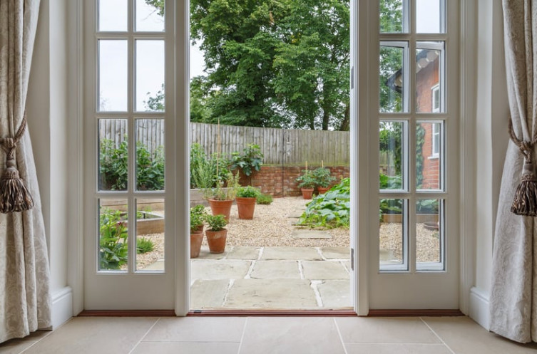 A sunlit open-concept living space with classic French doors leading to a garden patio, illustrating the benefits of installing French doors. The glass panels allow abundant natural light to flow in, creating a seamless indoor-outdoor connection. The elegant design enhances the home's aesthetic while improving ventilation and increasing property value.