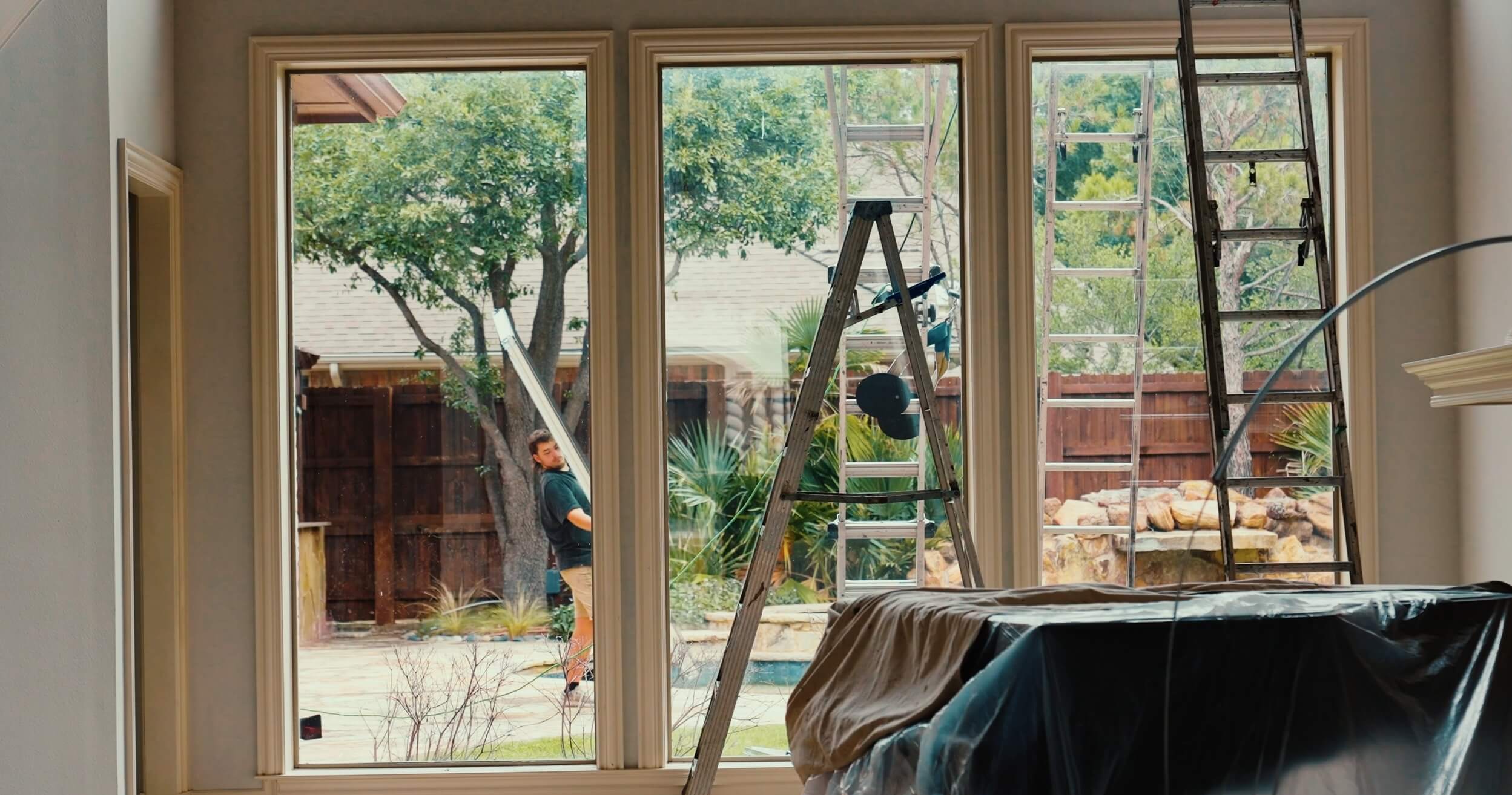 Three large windows show a view of a backyard with trees and a swimming pool. Two ladders are visible, one inside and one outside. A person stands outside near the window. Indoors, furniture is covered with a sheet.
