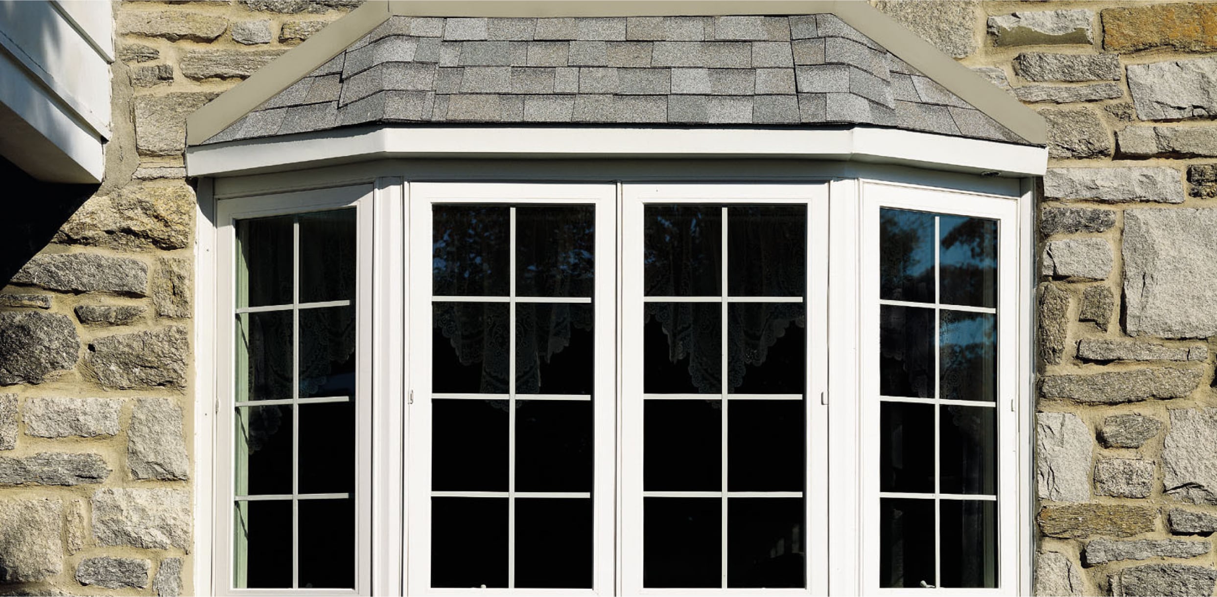 A white-framed bay window with multiple glass panes and grid patterns is installed in a stone exterior wall. The window has a shingled roof above it and lace curtains visible inside.