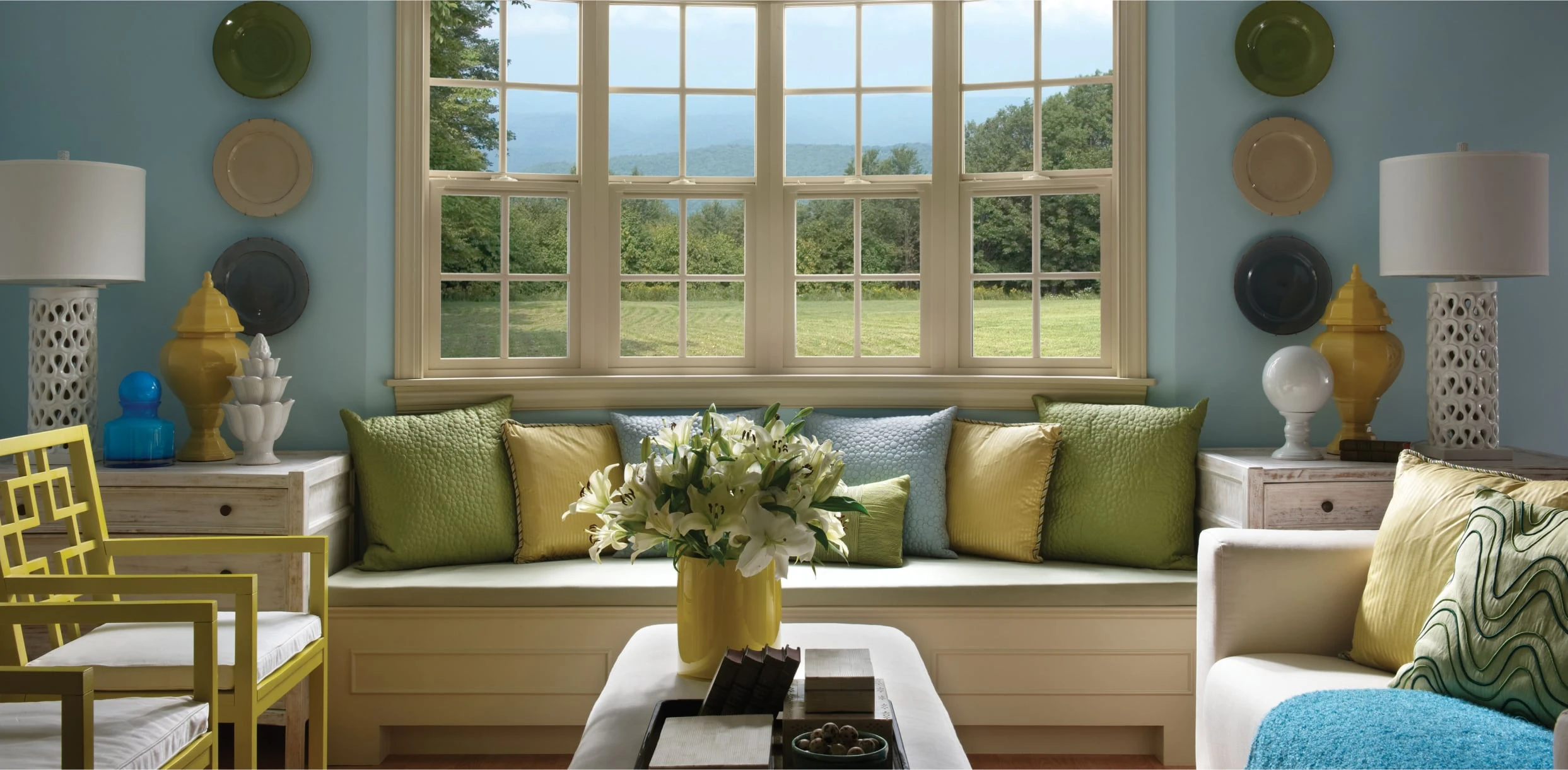 A bright living room with a large window showing a grassy field and trees, a built-in window seat with green and yellow cushions, white sofas, two table lamps, and a vase of white flowers on the coffee table.