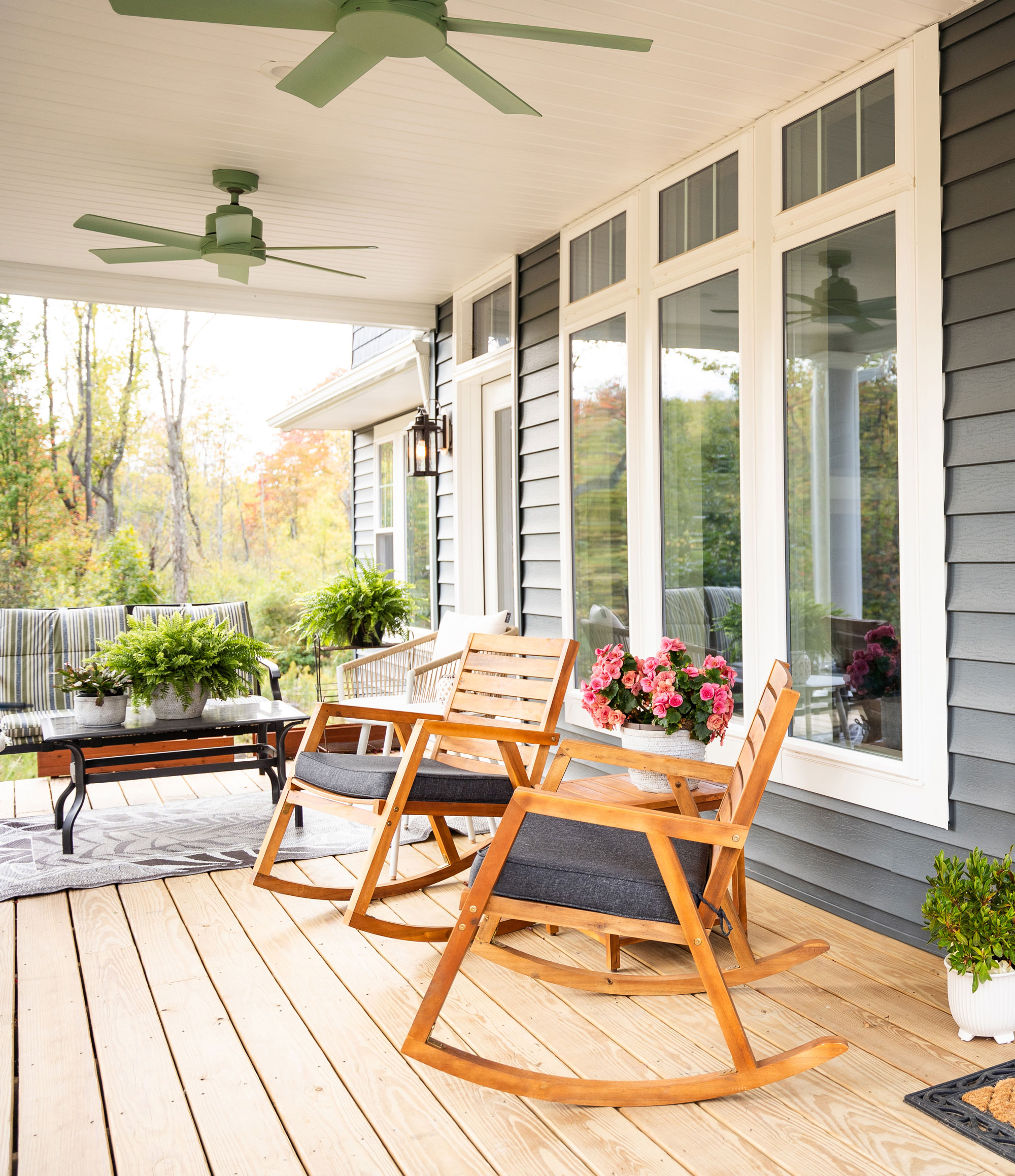 A wooden porch with two rocking chairs and a bench, potted plants, flowers, a small table, ceiling fans, and large windows on a gray house in a green, tree-filled setting.
