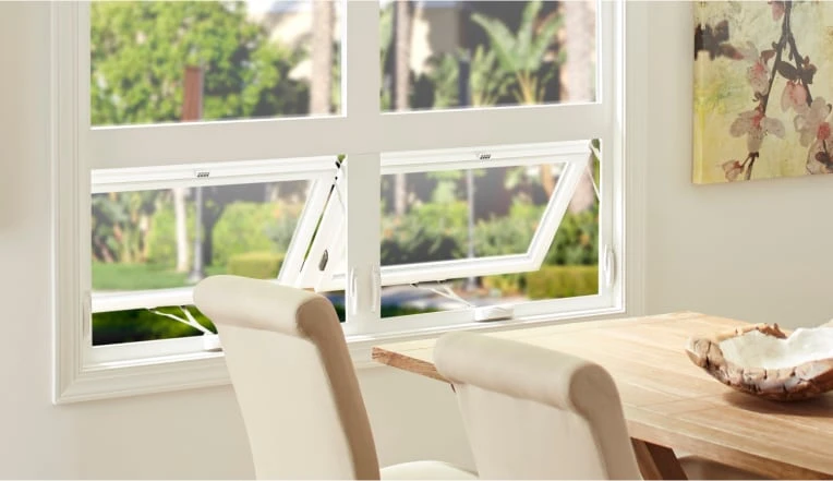 A dining area with cushioned chairs and a wooden table sits next to a large window with two lower panels open outward. Lush green plants and trees are visible outside. A floral painting hangs on the wall.