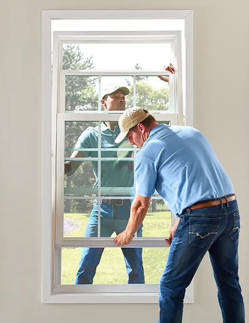 Two men installing a double-hung window; one man stands outside holding the window frame while the other man inside aligns and slides the window into place.