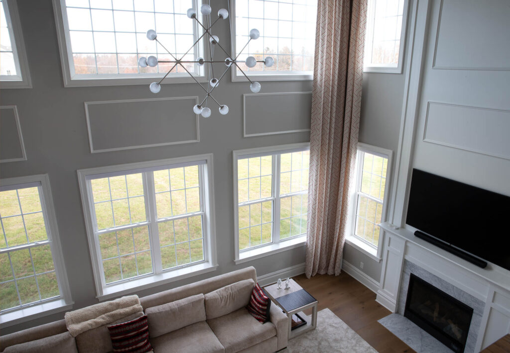 A modern living room with tall ceilings, large windows, a beige couch with red-striped pillows, a chandelier, a wall-mounted TV above a fireplace, and light brown curtains. Light streams in through the windows.