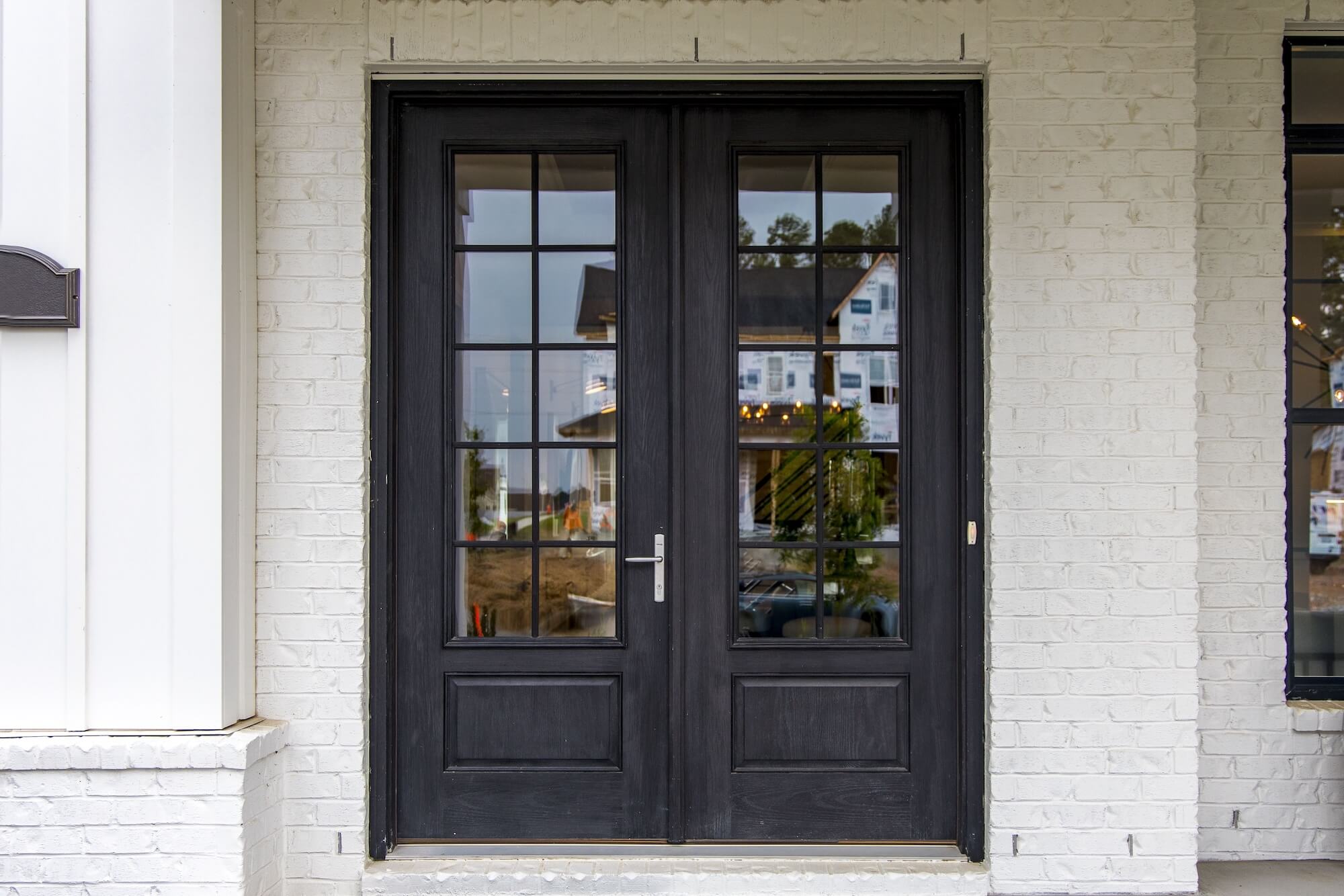 A set of double black framed glass doors with grid-style panes is set into a white painted brick wall. The reflection in the glass shows houses and trees across the street.