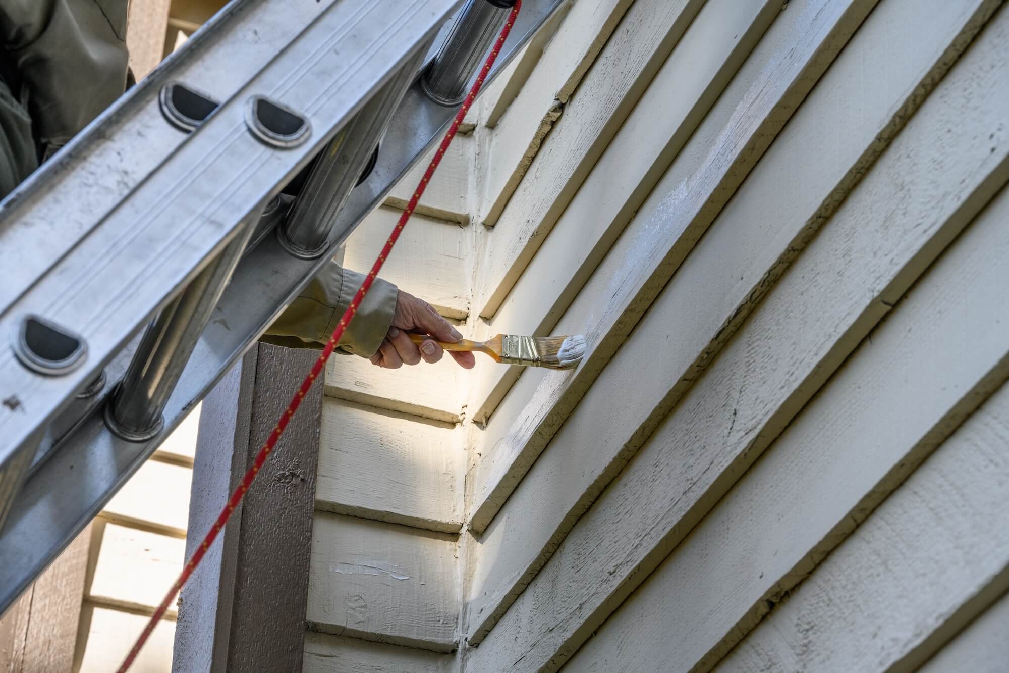 A person standing on a ladder uses a paintbrush to apply white paint to the exterior siding of a house. A red safety rope is attached to the ladder for stability.