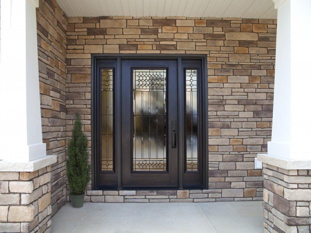 A modern front entrance with a black framed glass door, two side glass panels with decorative patterns, surrounded by light brown stone walls, a small potted tree on the left, and white columns.