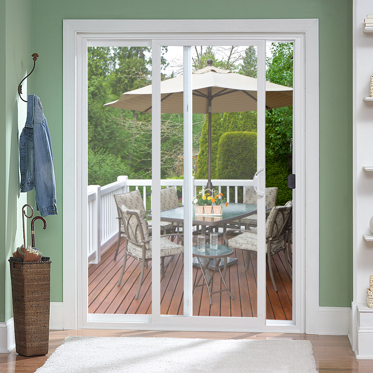A glass sliding door opens to a wooden deck with a round glass table, cushioned chairs, a vase of flowers, and a large patio umbrella. The indoor area has green walls, a white rug, and a coat rack with a jacket and umbrella.