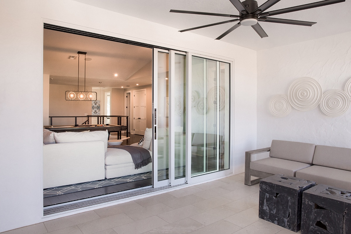 A modern indoor-outdoor space with a sliding glass door, a white sectional sofa and decorative lighting inside, and a gray outdoor sofa and two black cube tables on a light tile floor outside.