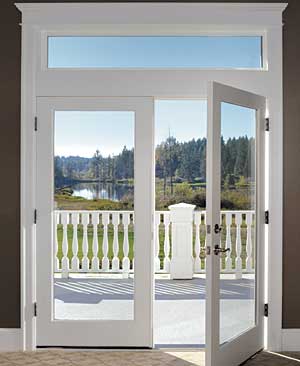 A white-framed glass door is open to a balcony with a white railing, overlooking a green landscape with trees and a body of water in the distance under a blue sky.