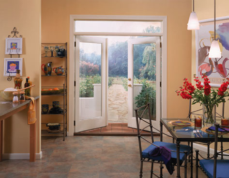A kitchen and dining area with a glass-top table, four chairs, red flowers in a vase, and a shelf with dishes. Double glass doors open to a stone path leading to a garden outside.