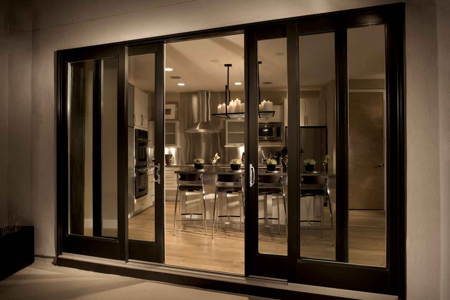 A view through large glass sliding doors into a modern kitchen and dining area, featuring a table with chairs, overhead lighting, stainless steel appliances, and candles on the table.