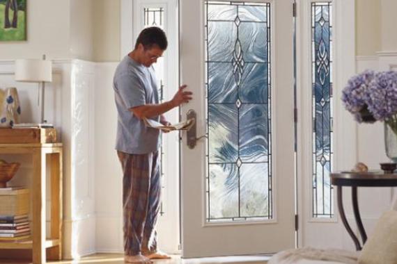 A man in pajamas stands by a glass-paneled front door, collecting mail from the mail slot. Sunlight comes through the door, and a table with books and a lamp is visible in the background.
