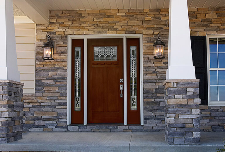 A front entryway with a wooden door, two sidelights featuring decorative glass, stone veneer walls, two wall-mounted lanterns, white columns, and part of a window to the right.