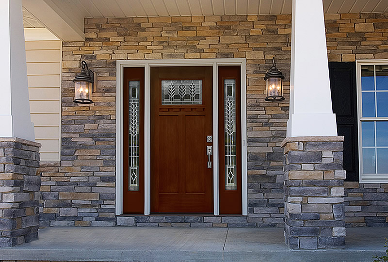 A front entryway with a wooden door, two sidelights featuring decorative glass, stone veneer walls, two wall-mounted lanterns, white columns, and part of a window to the right.